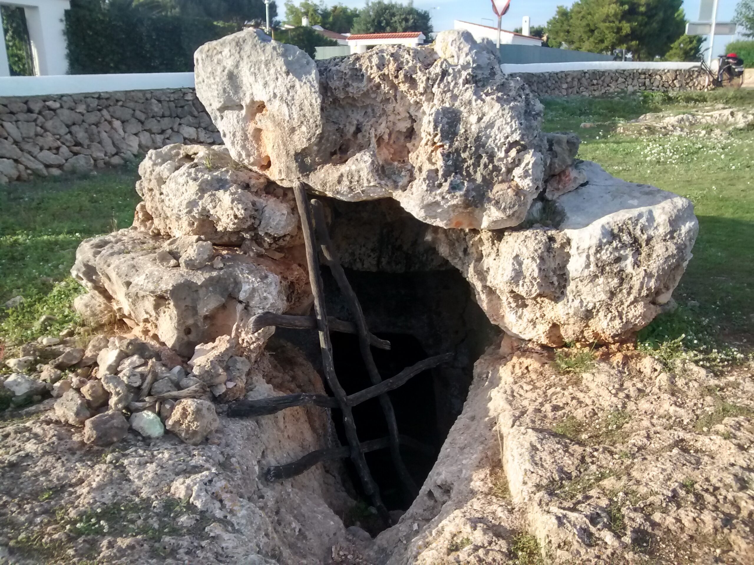Cala en Blanes Menorca: de kloofbaai bij Ciutadella, met Cala en Forcat en Los Delfines 3 Het hipogeu van Torre del Ram, een uitgehakte grafkamer uit de bronstijd met een ladder in de ingang, tussen gras en een droogstapelmuur