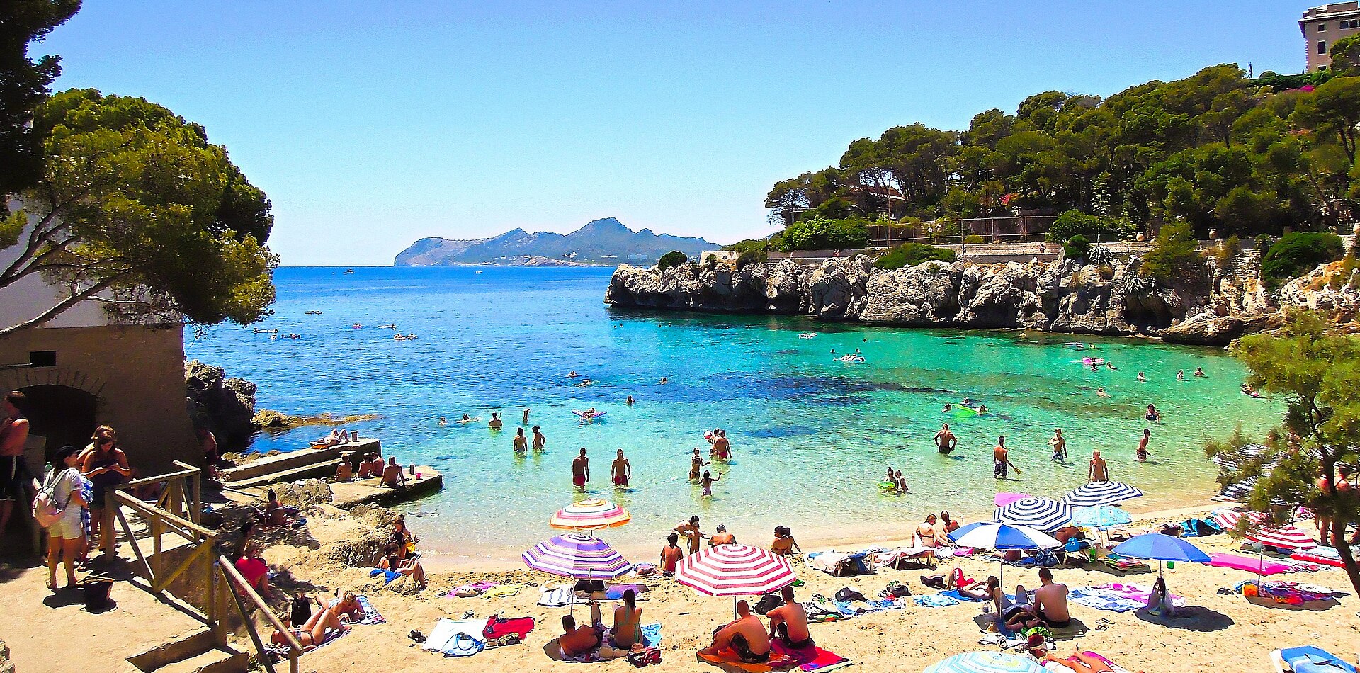 Cala Gat in de zomer met parasols op het zand, zwemmers in turquoise water en de bergen van de noordoostkust aan de horizon