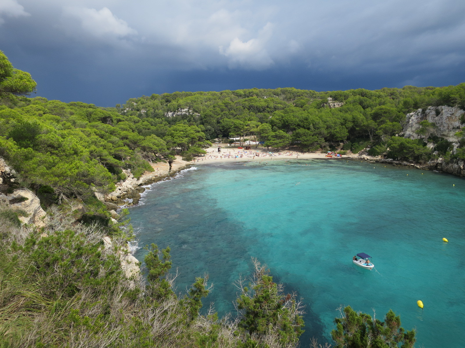 De baai van Cala Macarella onder donkere onweerswolken, met het strand en de strandtent in de verte
