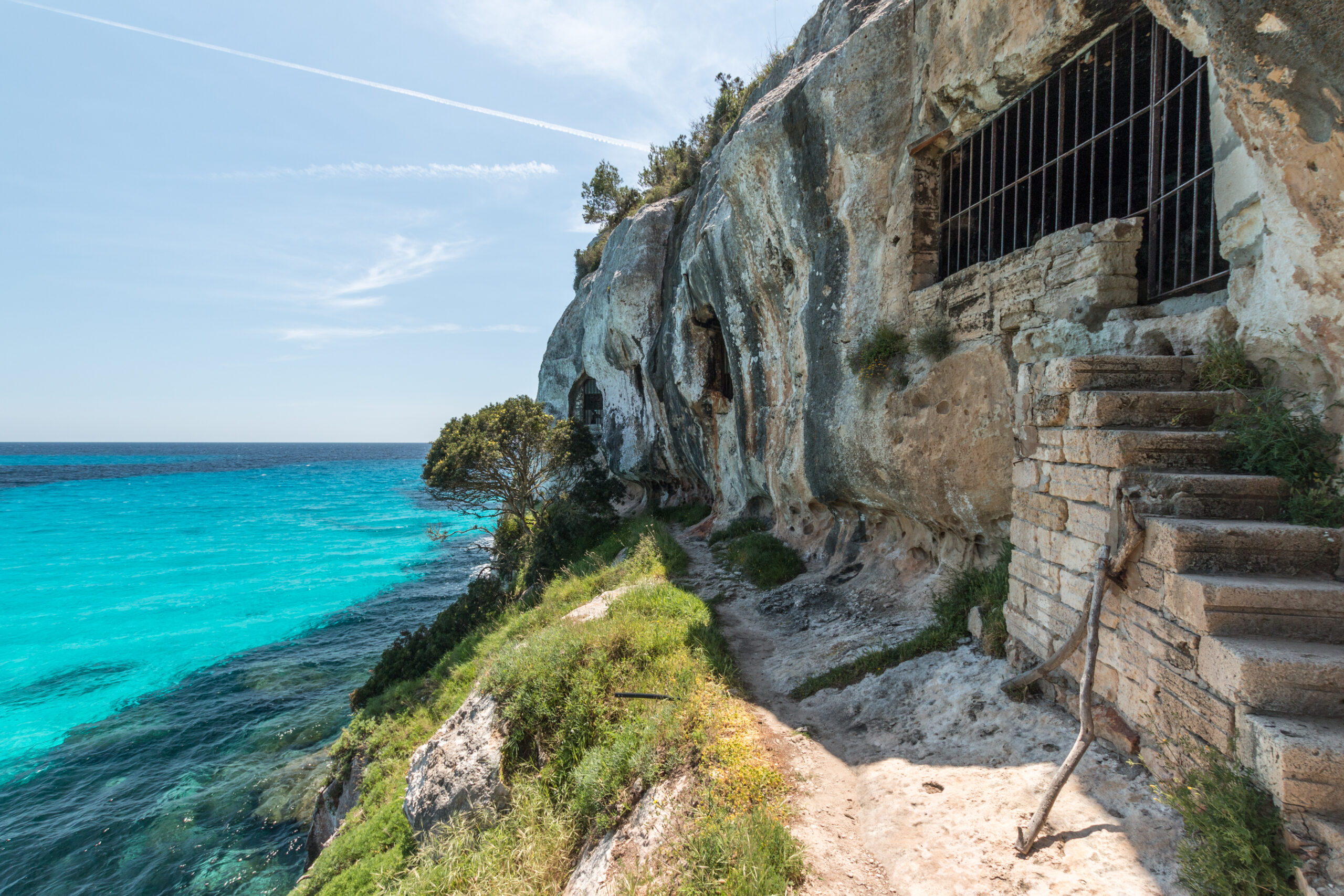 Het pad langs de kliffen bij Cala Macarella, met in de rots uitgehakte grotten en een getralied venster boven zee