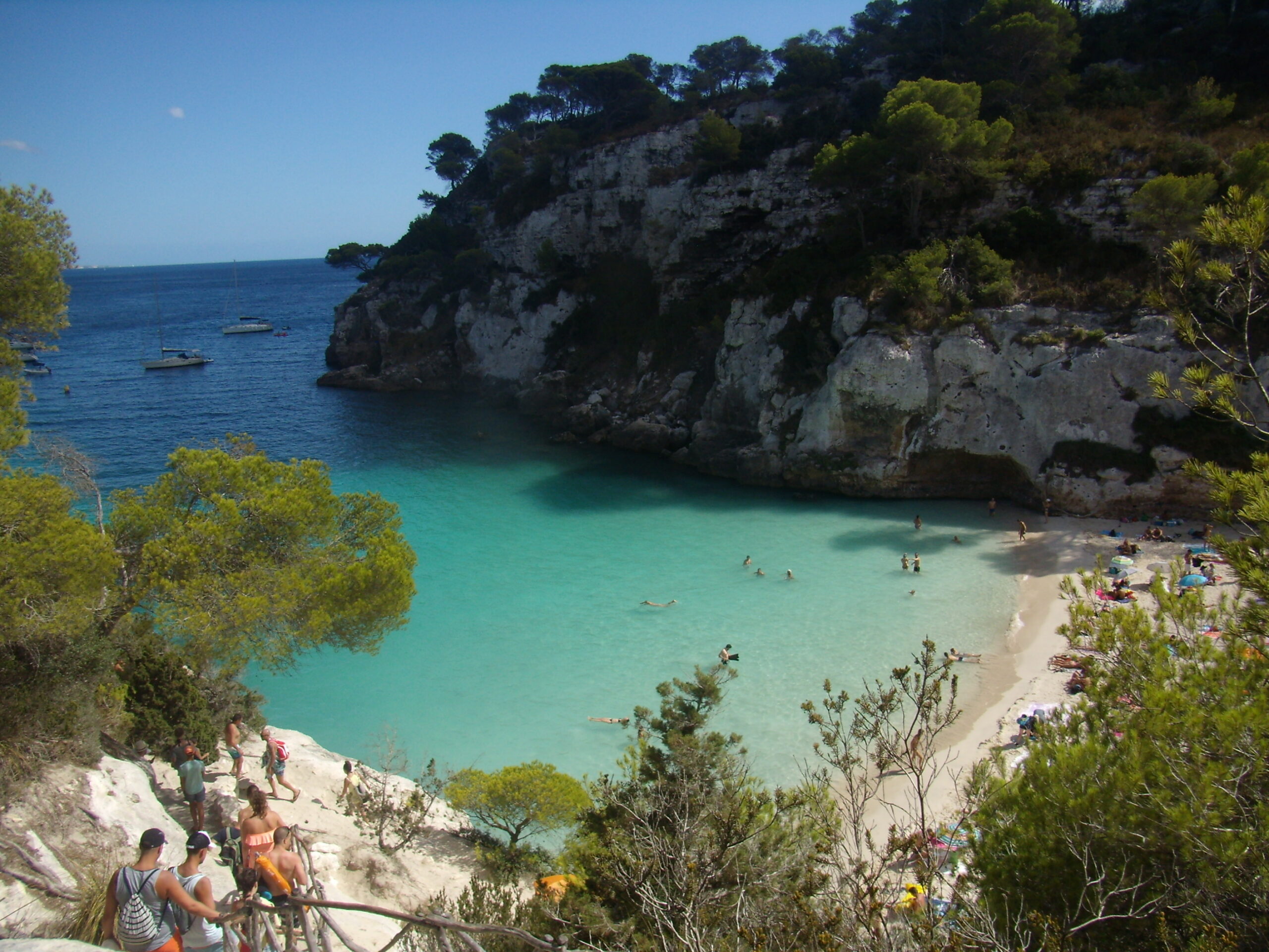 Cala Macarelleta van bovenaf, een klein wit zandstrand tussen de kliffen met zwemmers in het ondiepe water
