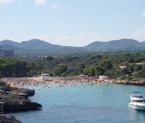 Cala Marçal van bovenaf gezien, met rijen rieten parasols op het zand, zwemmers in turquoise water, een motorjacht in de baai en de heuvels van het achterland
