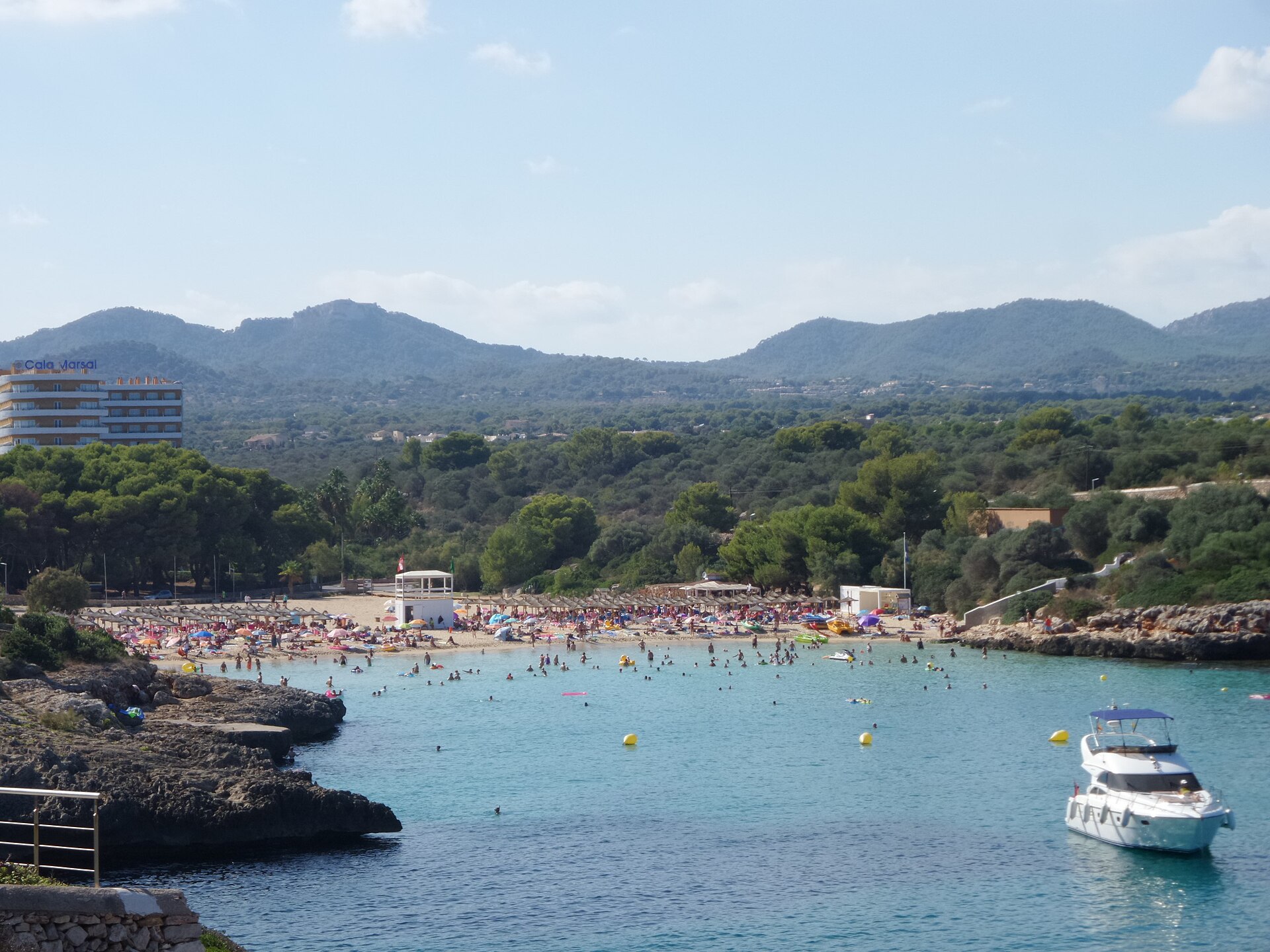 Cala Marçal van bovenaf gezien, met rijen rieten parasols op het zand, zwemmers in turquoise water, een motorjacht in de baai en de heuvels van het achterland