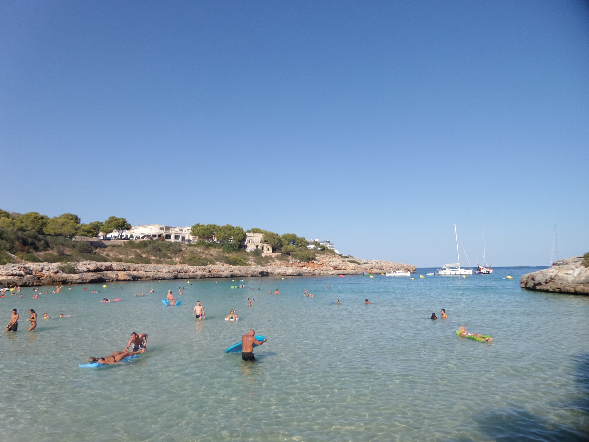Vanuit het water gezien bij Cala Marçal: zwemmers en luchtbedden boven een zandbodem, met de lage rotsrichels en zeilboten bij de monding van de baai