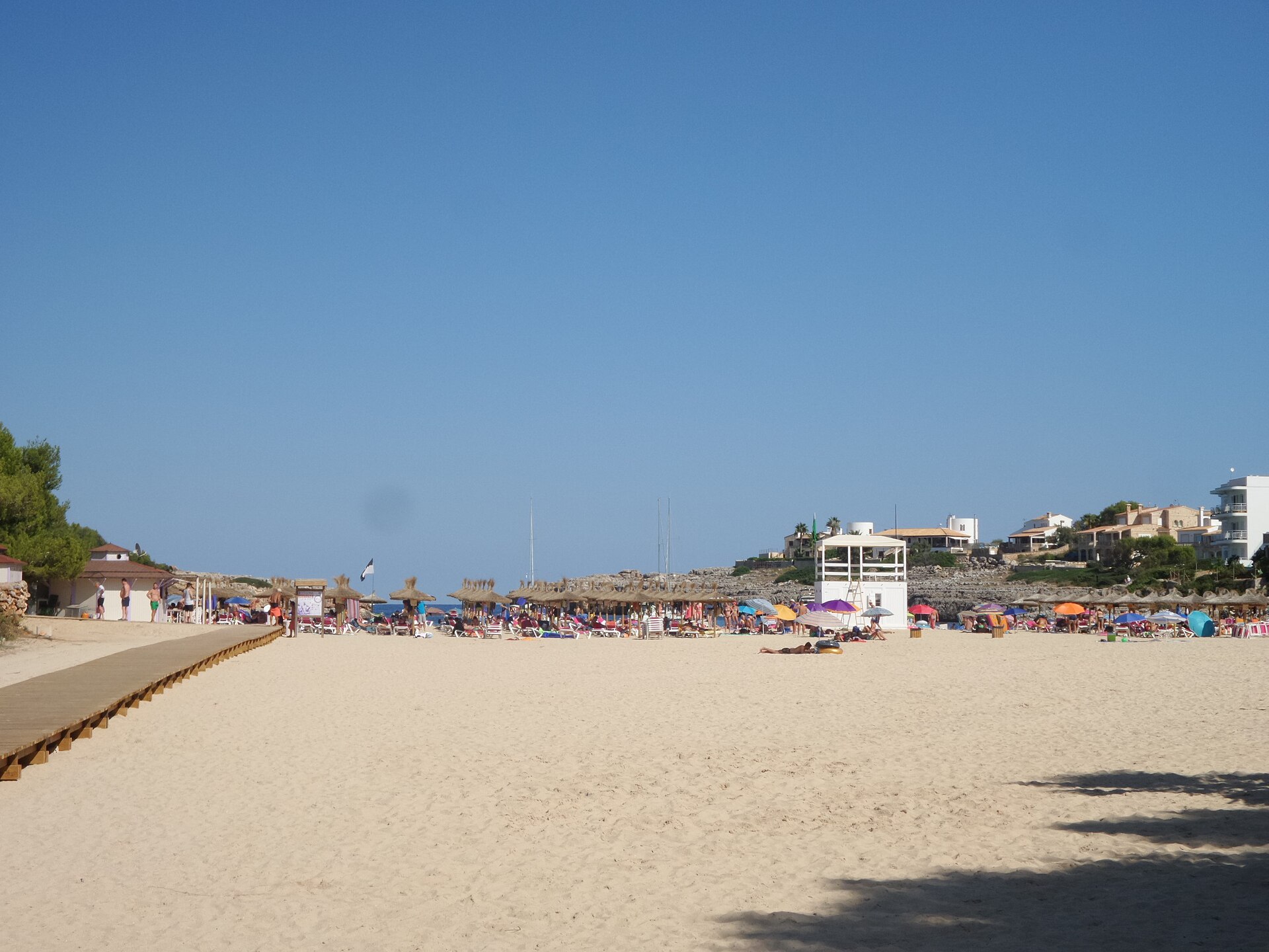 Het brede zandstrand van Cala Marçal met badgasten, parasols en de witte uitkijkpost van de strandwacht