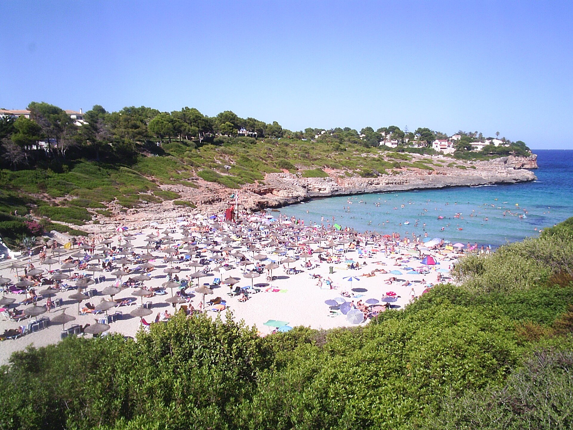 Cala Mendia van bovenaf in het hoogseizoen, met het zand helemaal vol parasols en handdoeken en de huizen rond de baai