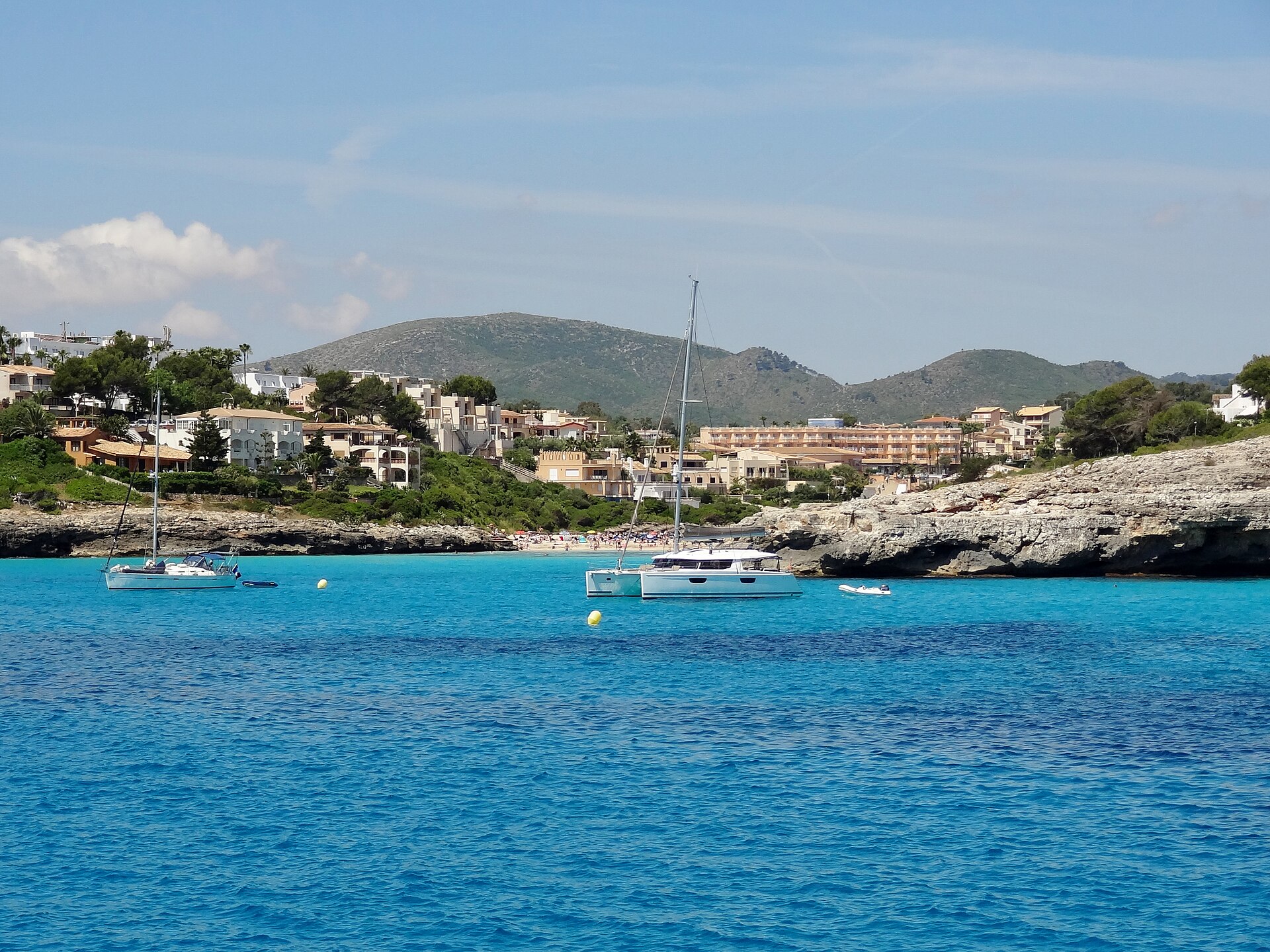 De monding van Cala Mendia met ankerende zeilboten op turquoise water en de bebouwing en heuvels aan de kust erachter