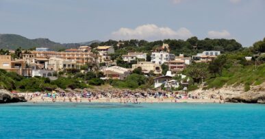 Cala Mendia vanaf zee gezien, met een rij rieten parasols op het witte zand, badgasten in turquoise water en het dorp dat tegen de beboste helling op loopt