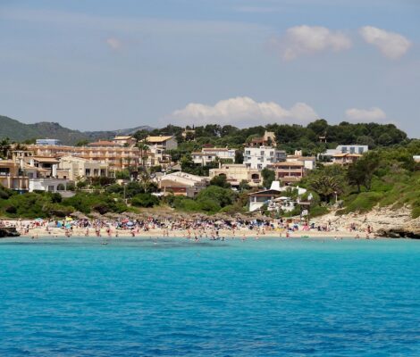 Cala Mendia vanaf zee gezien, met een rij rieten parasols op het witte zand, badgasten in turquoise water en het dorp dat tegen de beboste helling op loopt