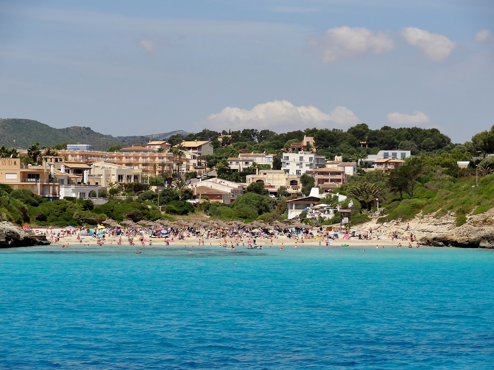 Cala Mendia vanaf zee gezien, met een rij rieten parasols op het witte zand, badgasten in turquoise water en het dorp dat tegen de beboste helling op loopt