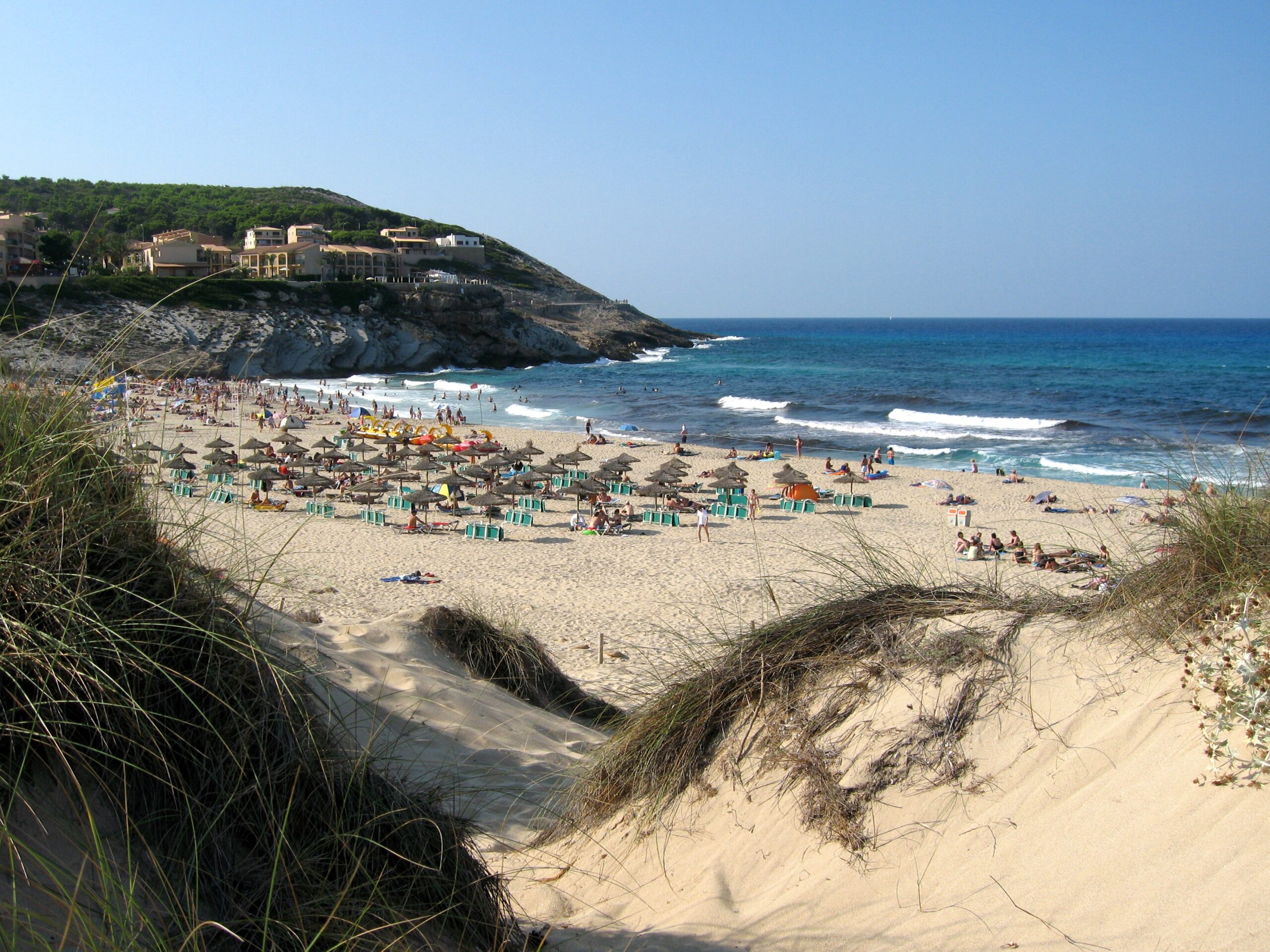 Uitzicht over een duinrug met helmgras op het volle zomerstrand van Cala Mesquida, met rijen rieten parasols en de hotels op de landtong erachter