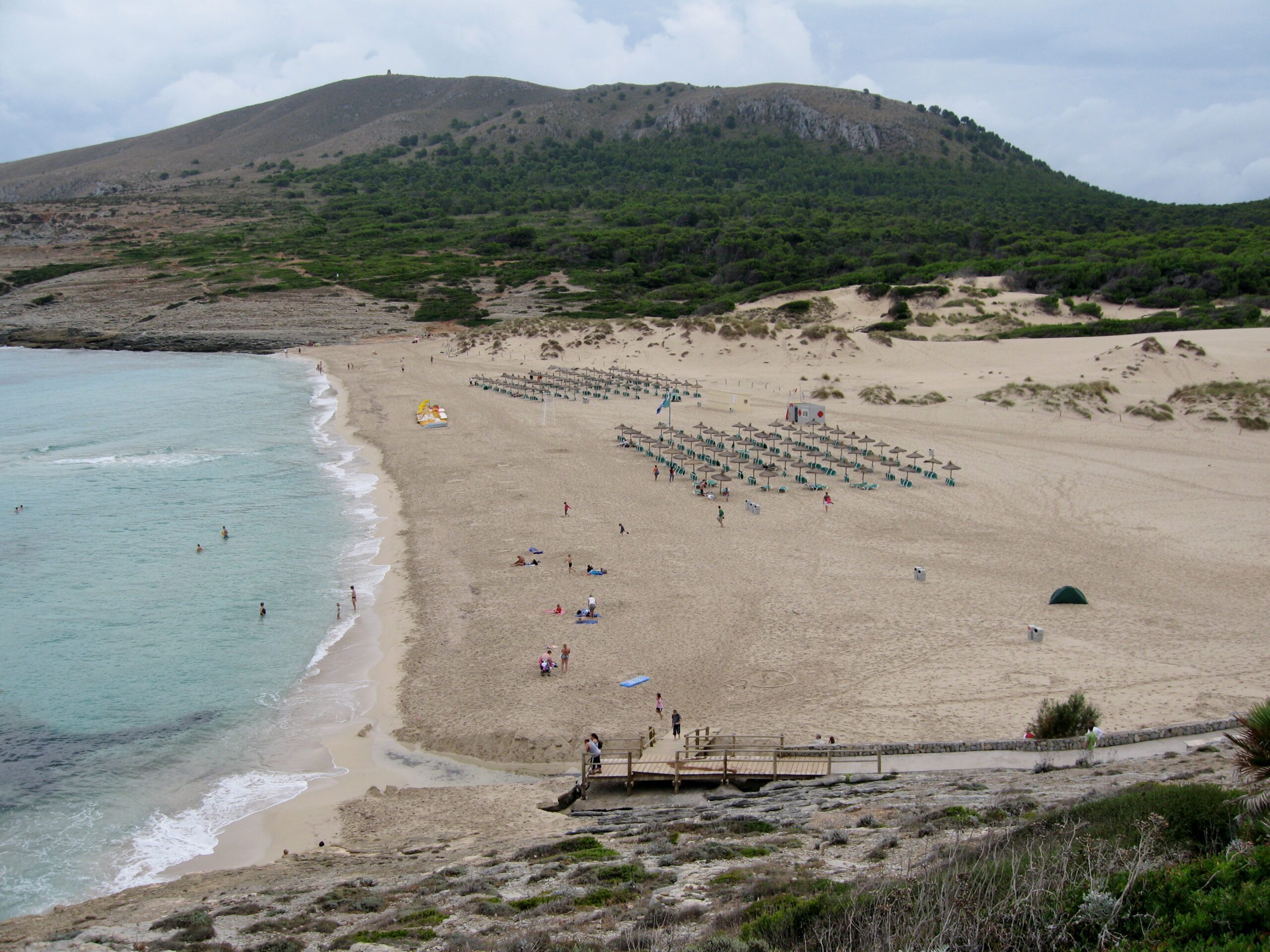 Het brede zandstrand van Cala Mesquida met strakke rijen parasols, het houten vlonderpad naar de duinen en de kale heuvel van de Talaia erachter