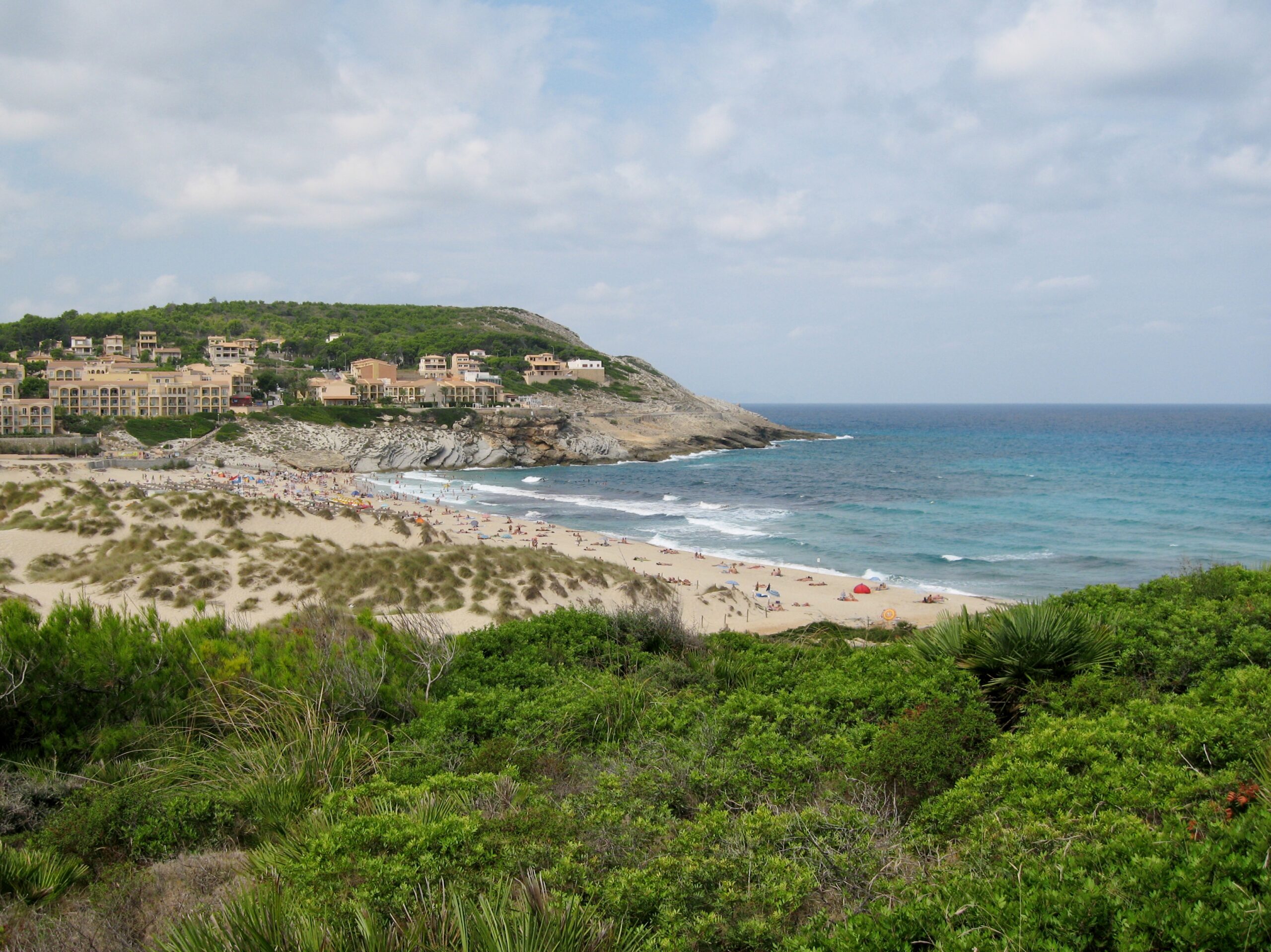 De baai van Cala Mesquida gezien over de duinen en de lage struiken, met het vakantiedorp op de rotsige landtong aan de linkerkant