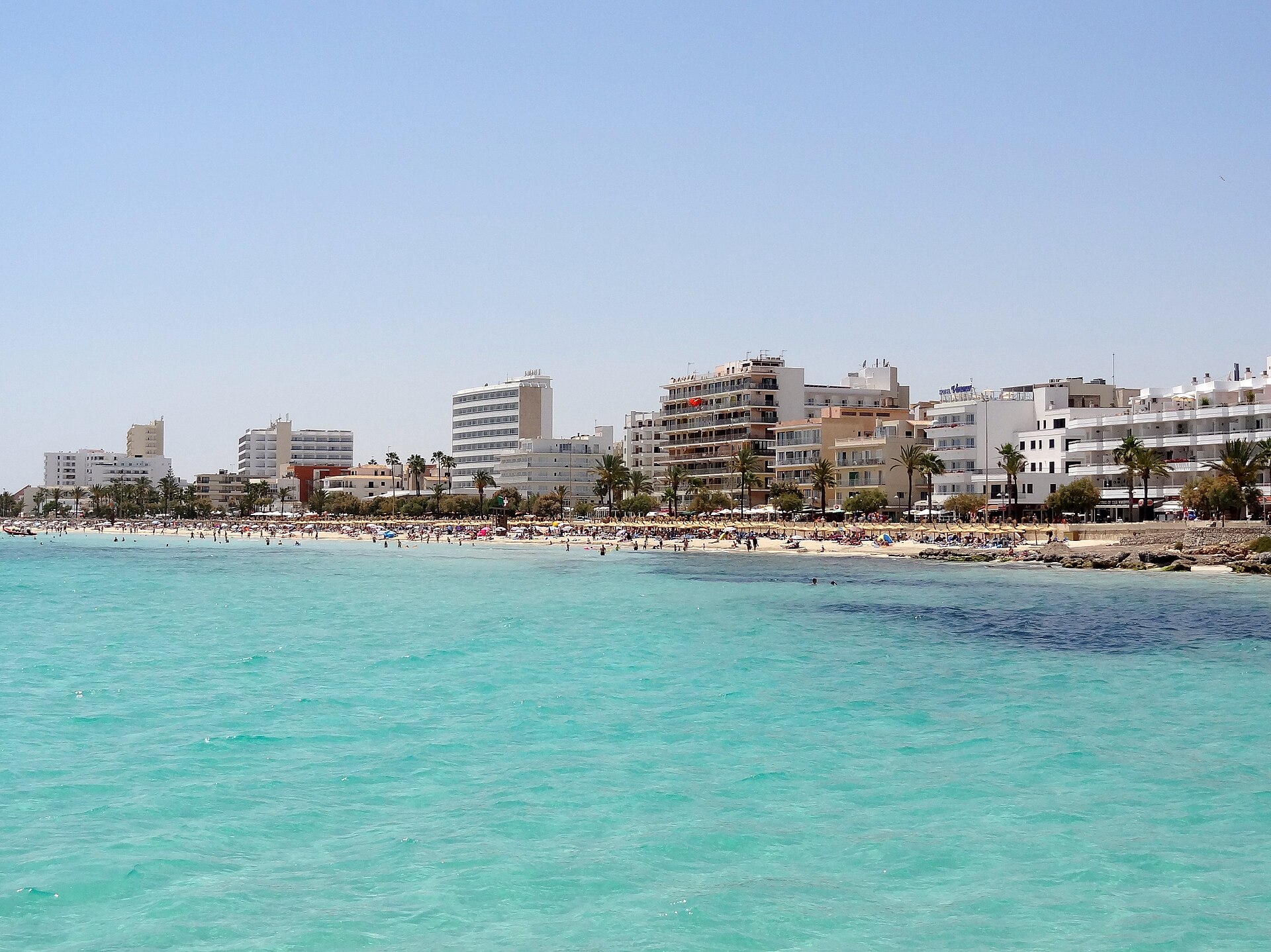 Helder turquoise water voor het strand van Cala Millor, met de lange rij hotels en palmen aan de overkant van de baai