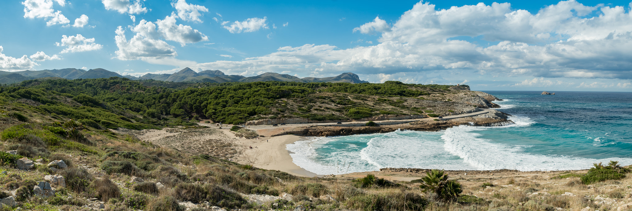 Cala Mitjana Mallorca bij Artà: wilde baai, wandeling en parkeren 2 Cala Mitjana bij Arta vanaf de heuvel erboven, met witte branding in de baai, struikgewas en pijnbos eromheen en de bergen van het schiereiland op de achtergrond