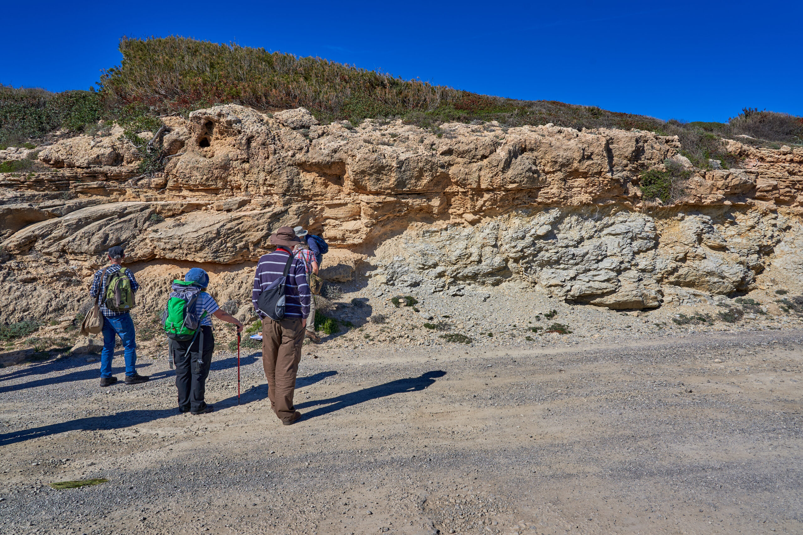 Cala Mitjana Mallorca bij Artà: wilde baai, wandeling en parkeren 5 Wandelaars op het grindpad naar Cala Mitjana, voor een rotswand waarin lichtbruine kalkzandsteen van een fossiel duin op veel oudere, sterk gebroken kalksteen ligt