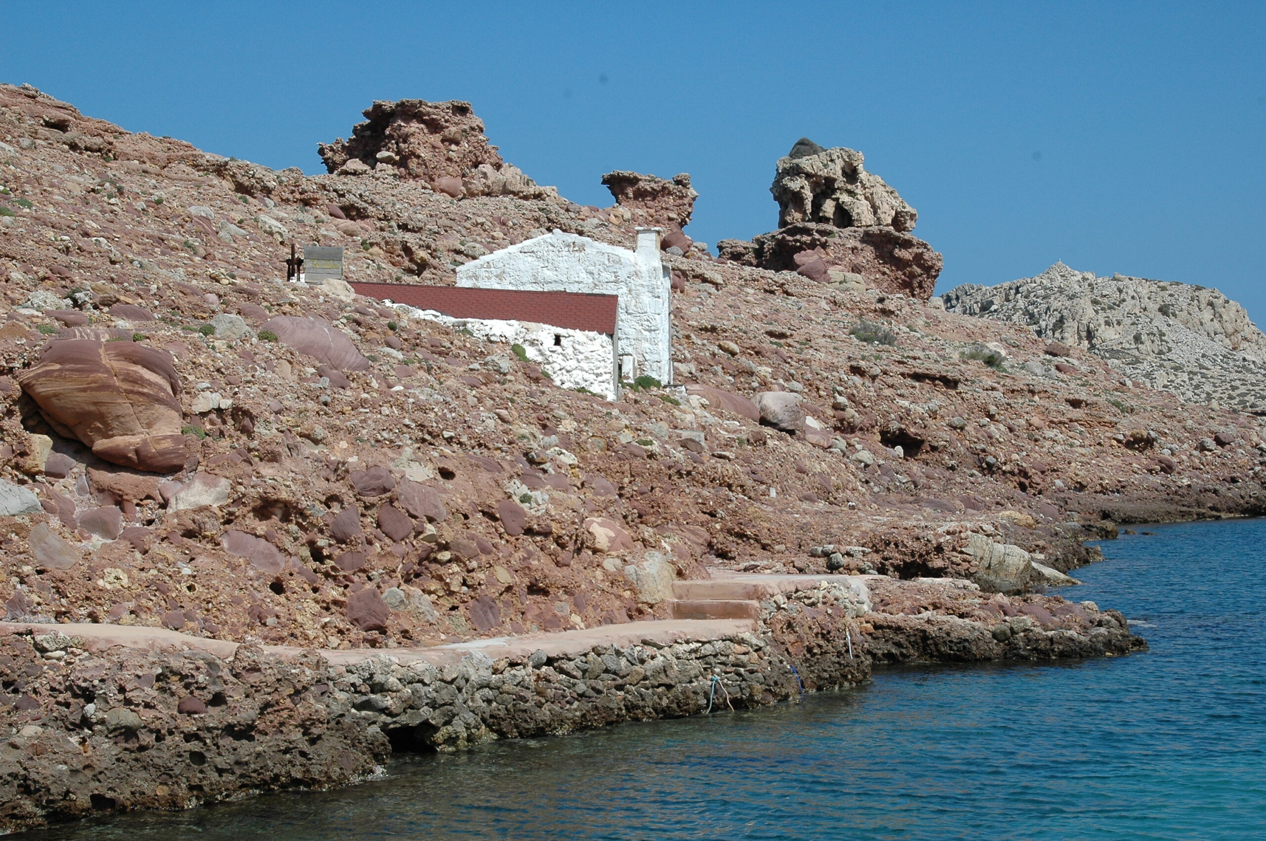 Cala Morell Menorca: witte huizen, grafgrotten en een klein rotsstrand 6 Wit bootshuis tegen de rode conglomeraatrotsen van Cala Morell, met betonnen terrassen aan het water