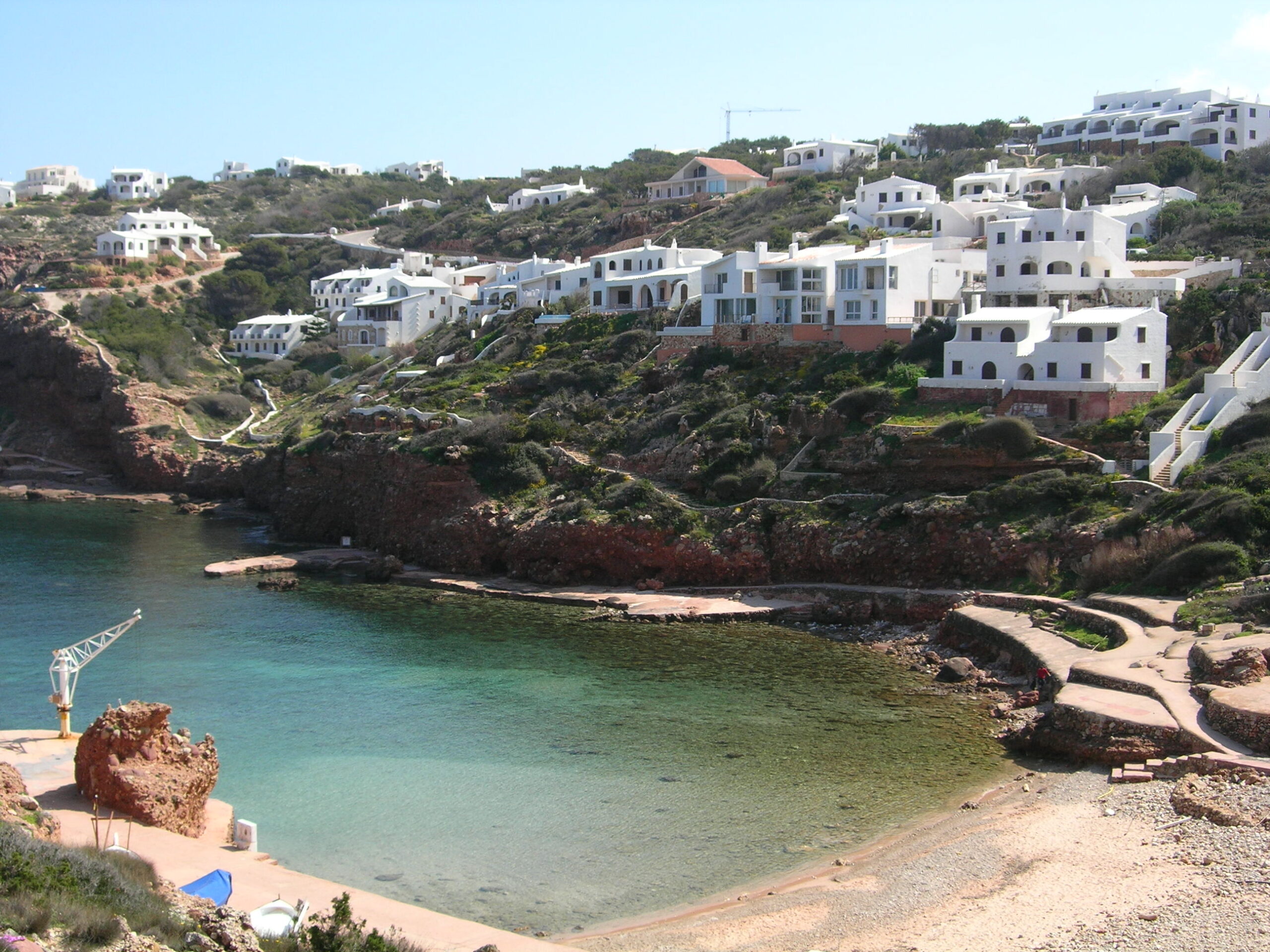 Cala Morell Menorca: witte huizen, grafgrotten en een klein rotsstrand 3 Het kleine kiezelstrand van Cala Morell met betonnen zonneterrassen op de rode rotsen en witte huizen erboven