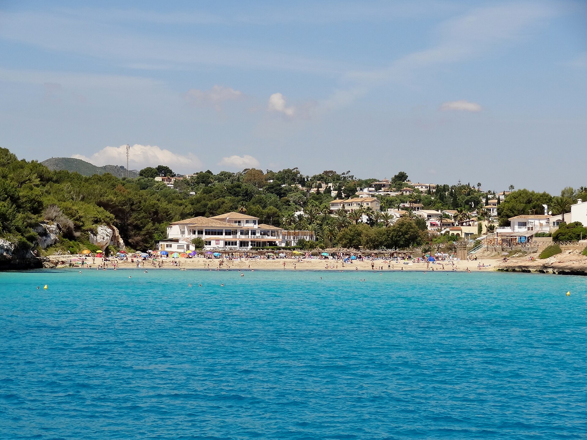 Cala Romàntica vanaf zee gezien, met het lichte zandstrand, de bebouwing erachter en de beboste helling
