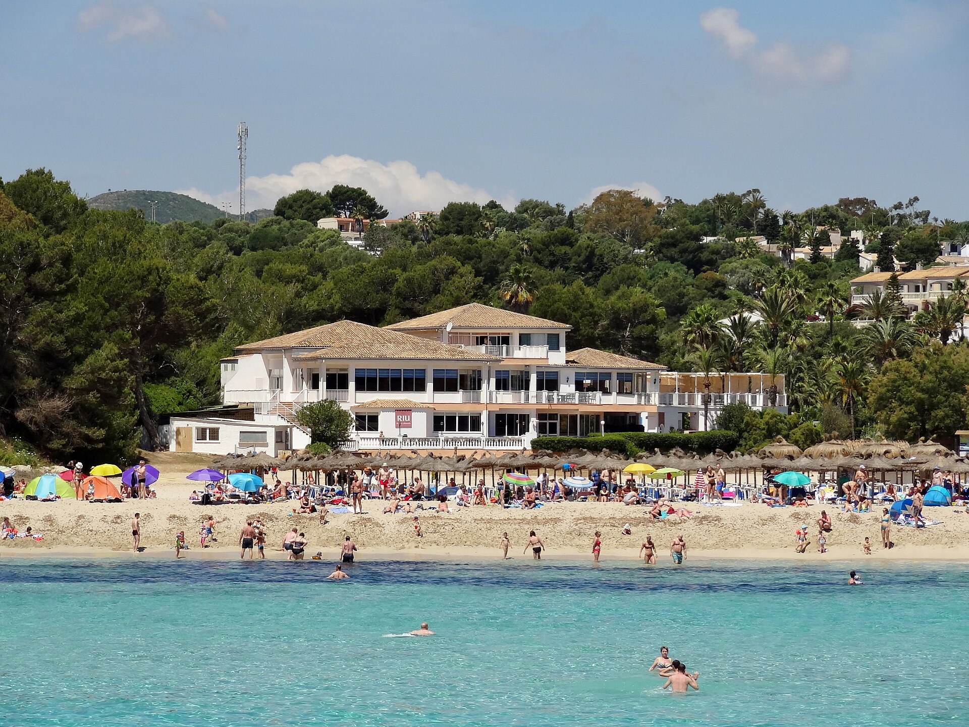 Het strand van Cala Romàntica in het hoogseizoen, met een lange rij rieten parasols, badgasten op het lichte zand, turquoise water en het hotel en de pijnbomen erachter