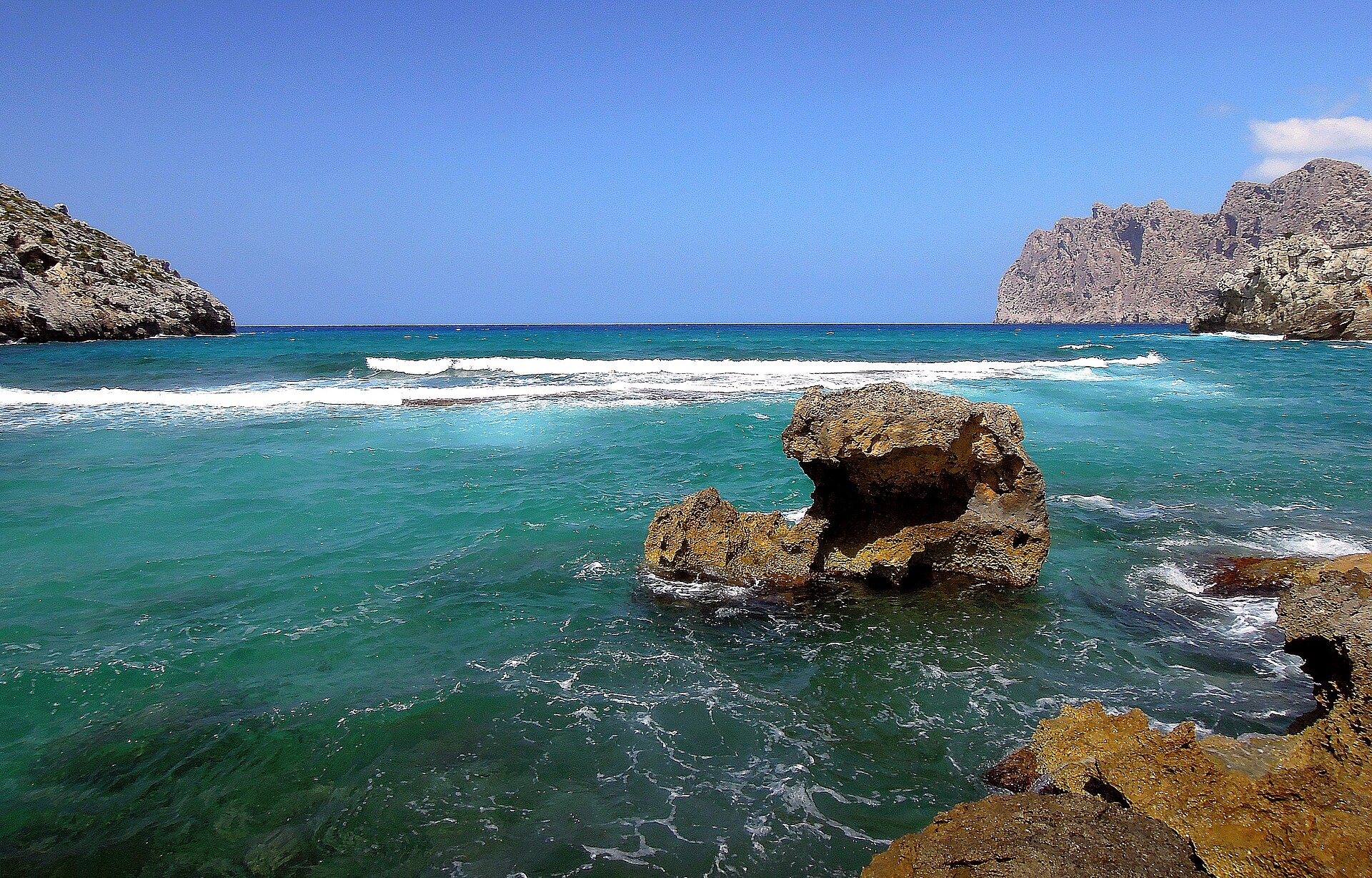 Turquoise water en een losse rots voor de kust bij Cala Clara, met de steile kliffen aan de overkant van de baai
