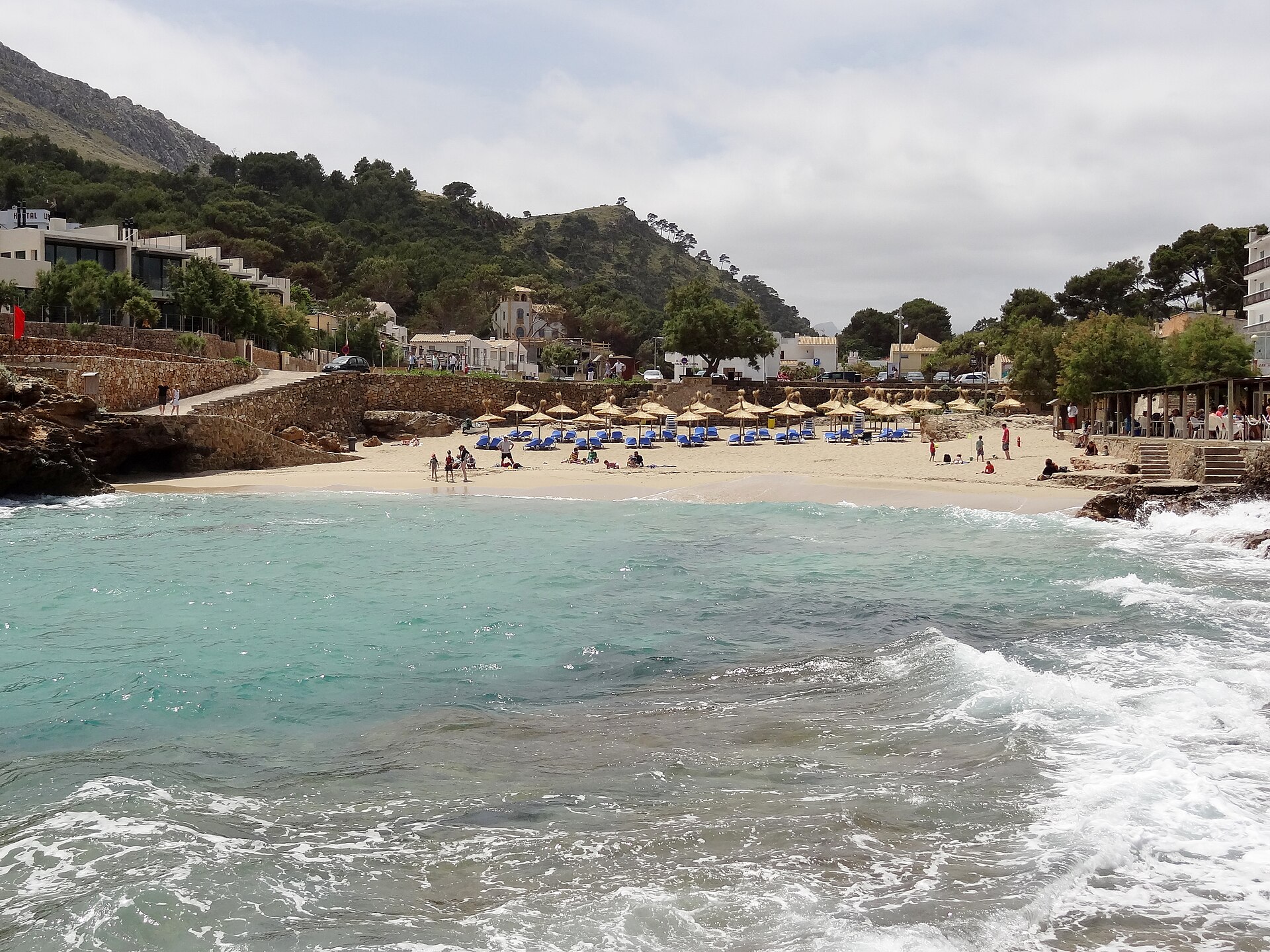 Het brede zandstrand van Cala Molins met rieten parasols, de huizen van het dorp en branding op de voorgrond