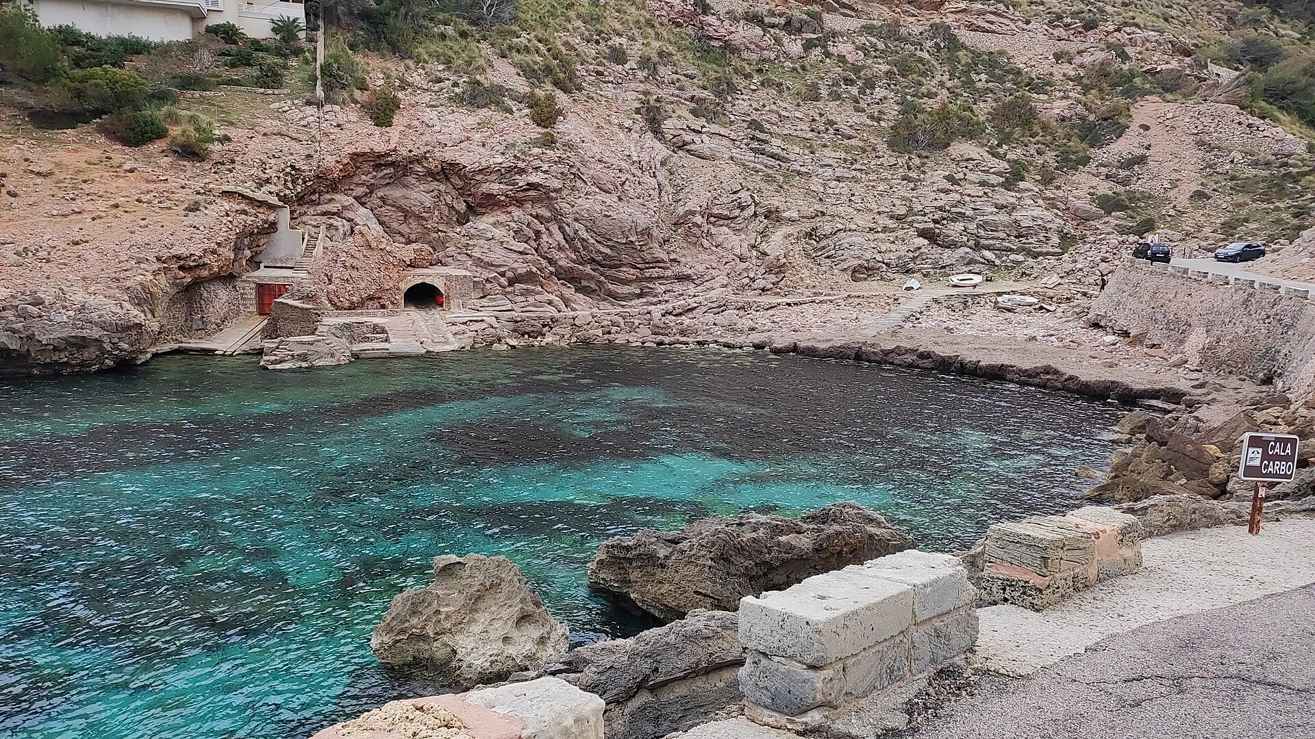 Het rotsachtige baaitje Cala Carbó met helder groenblauw water, botenhuizen in de rotswand en het bordje met de naam van de cala