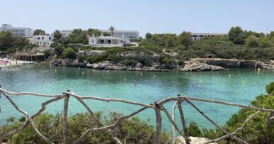 Cala Santandría Menorca: de beschutte kreek bij Ciutadella en de Torre des Castellar 13 Het turquoise water van Cala Santandría op Menorca, gezien over een houten hek met de witte huizen op de rotsrand erachter