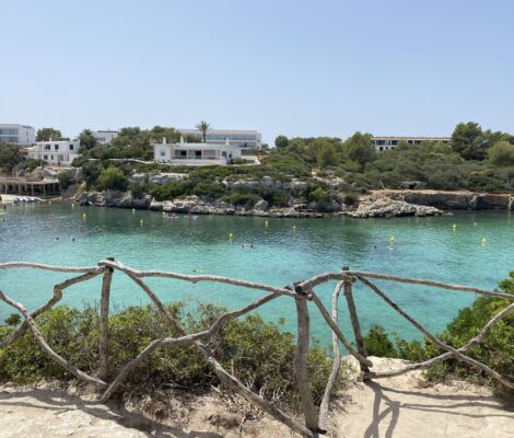 Het turquoise water van Cala Santandría op Menorca, gezien over een houten hek met de witte huizen op de rotsrand erachter