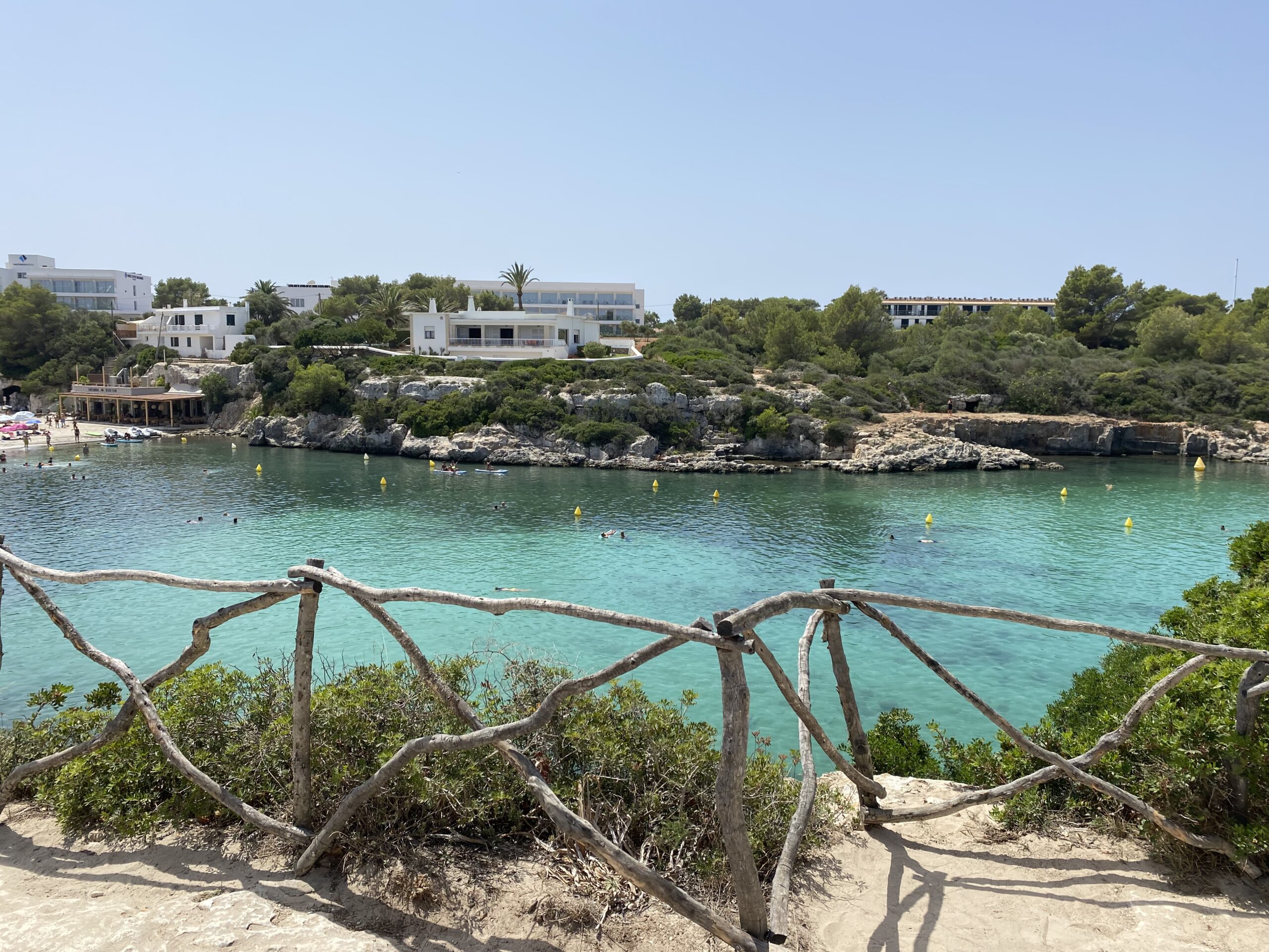 Cala Santandría Menorca: de beschutte kreek bij Ciutadella en de Torre des Castellar 1 Het turquoise water van Cala Santandría op Menorca, gezien over een houten hek met de witte huizen op de rotsrand erachter