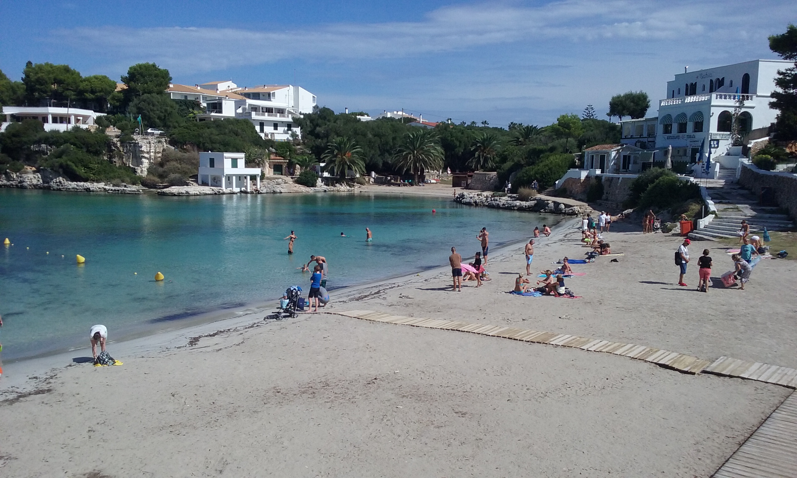 Cala Santandría Menorca: de beschutte kreek bij Ciutadella en de Torre des Castellar 4 De zandstrook achterin Cala Santandría met badgasten, vlonderpad en het witte gebouw met boogramen aan de rand van de inham