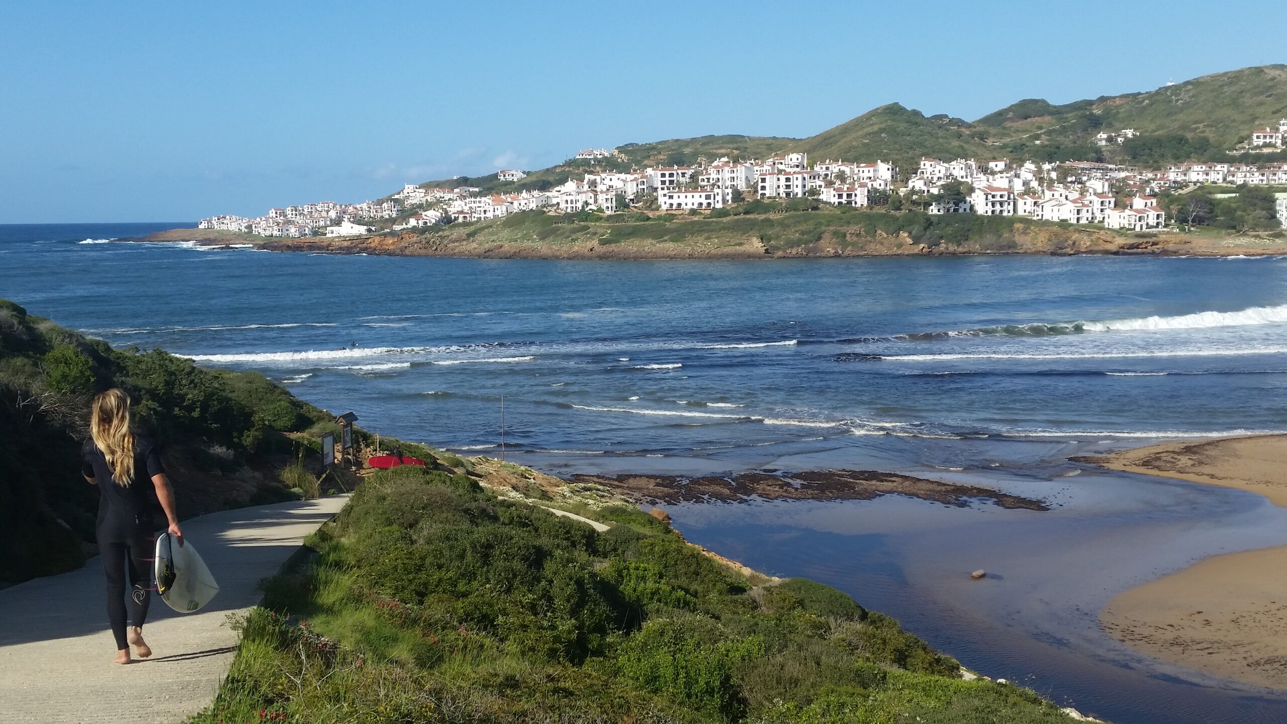 Een surfer loopt met zijn plank naar Cala Tirant terwijl golven de baai in rollen