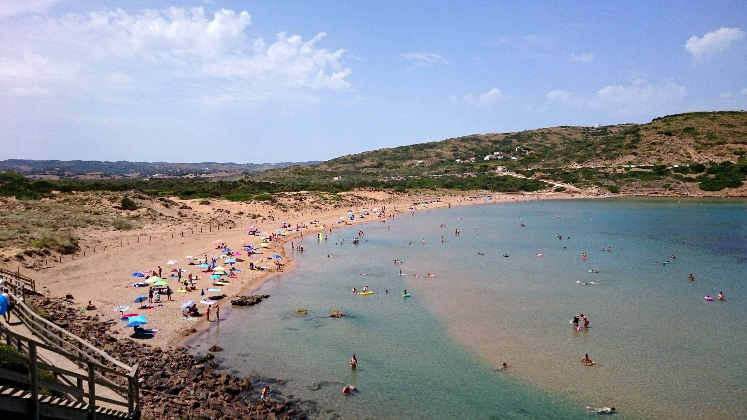 De zandboog van Cala Tirant in augustus met parasols, ondiep turquoise water en de duinen erachter