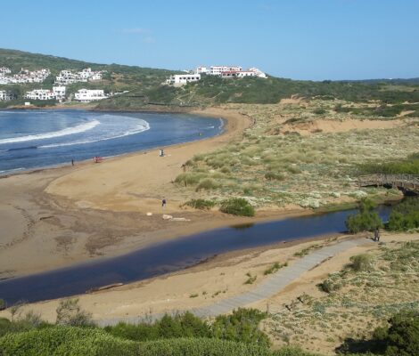 Het brede strand van Cala Tirant op Menorca met het beekje, de houten brug over het moeras en de witte huizen van Platges de Fornells