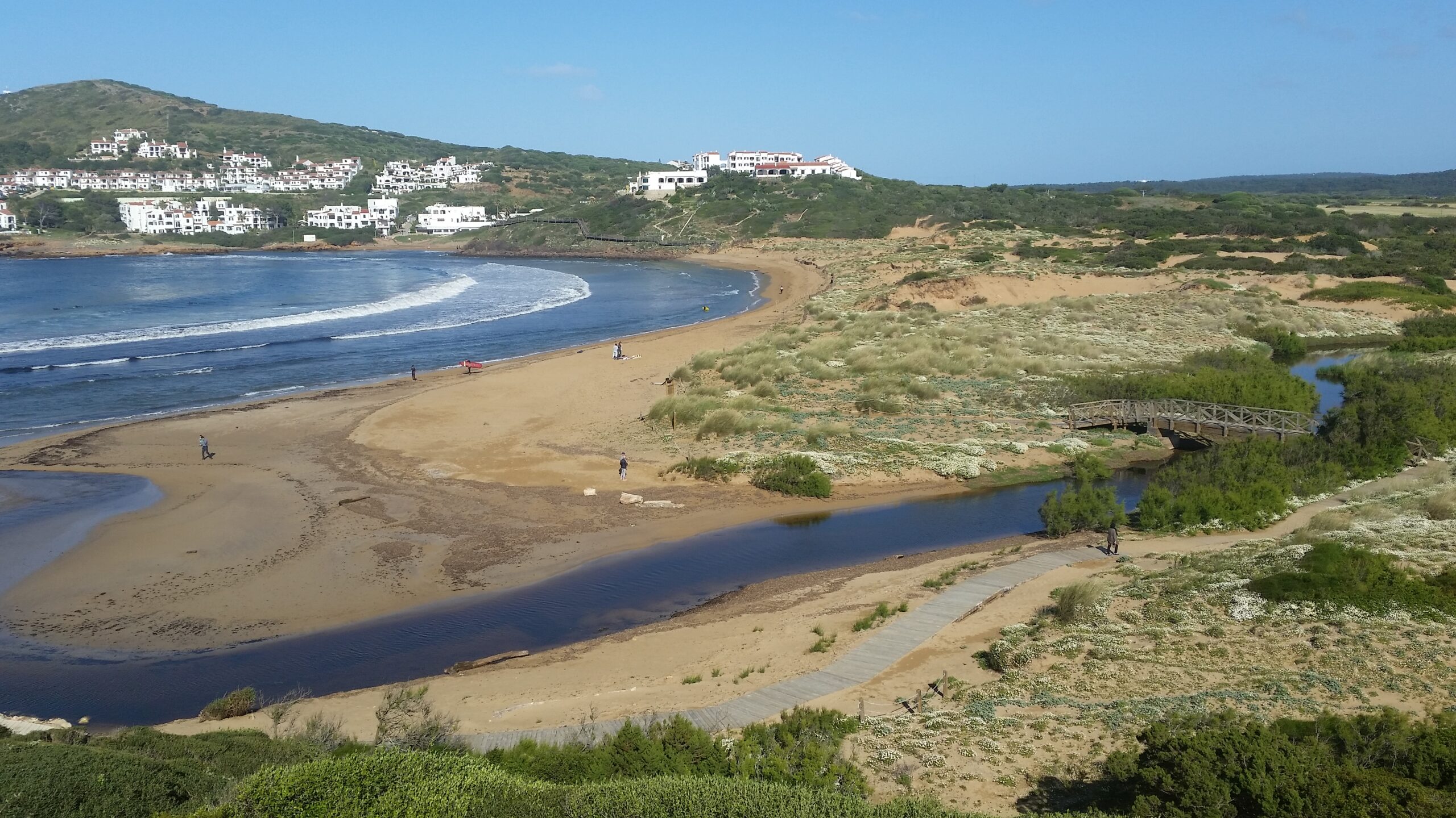 Het brede strand van Cala Tirant op Menorca met het beekje, de houten brug over het moeras en de witte huizen van Platges de Fornells