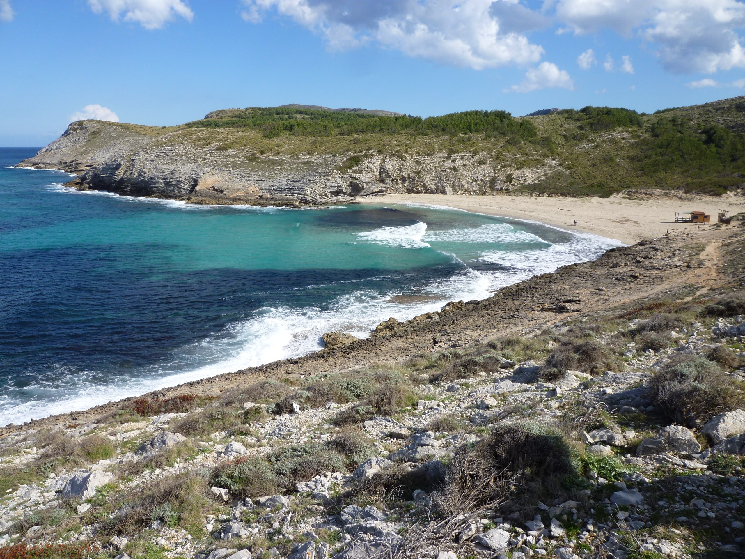 Het strand van Cala Torta gezien vanaf de rotsen aan de zuidkant, met brekende golven, turquoise water en onbebouwde heuvels rondom de baai