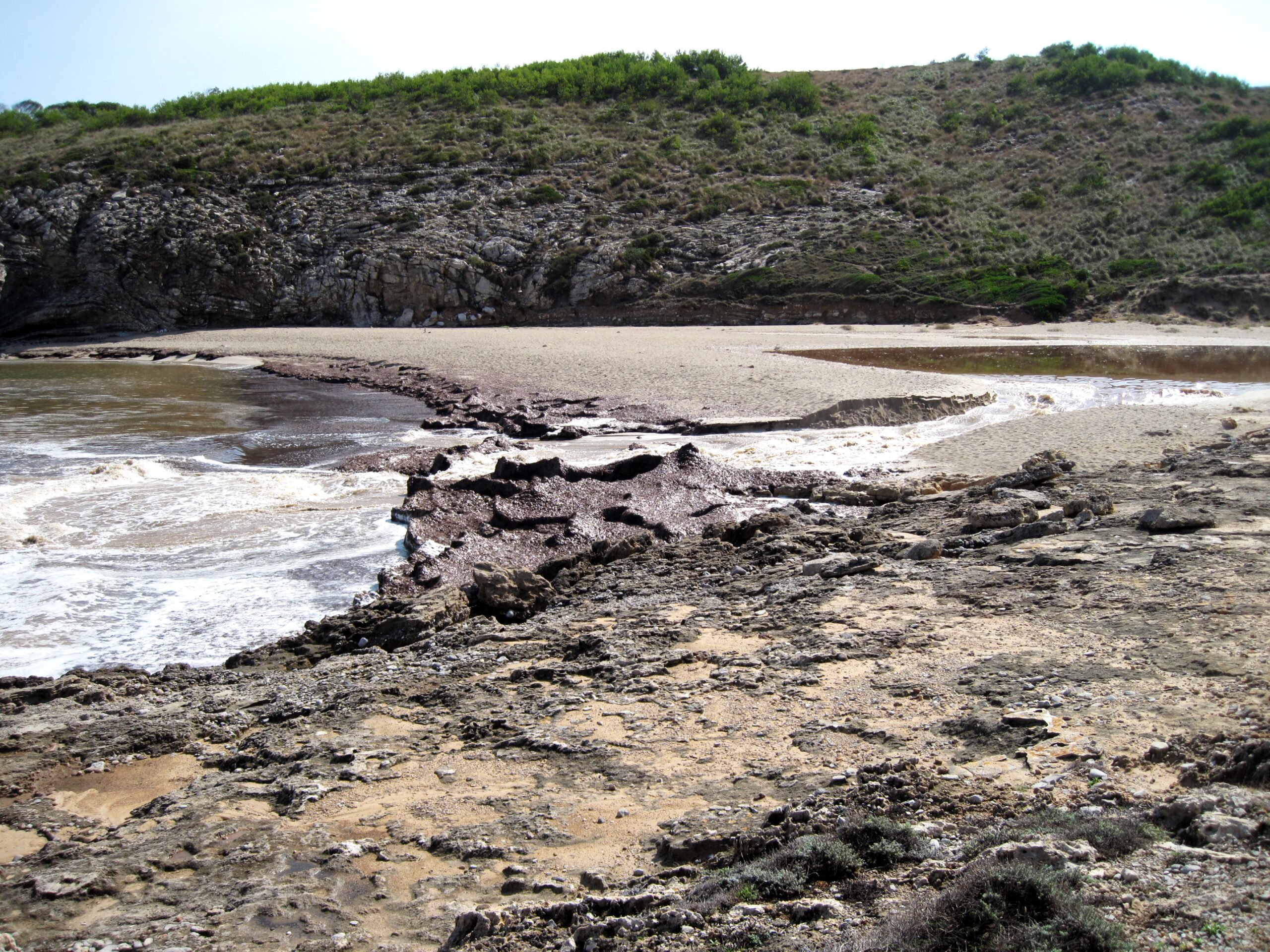 Golven die over de vlakke rotsrand aan de rand van Cala Torta slaan, met het lege zandstrand erachter