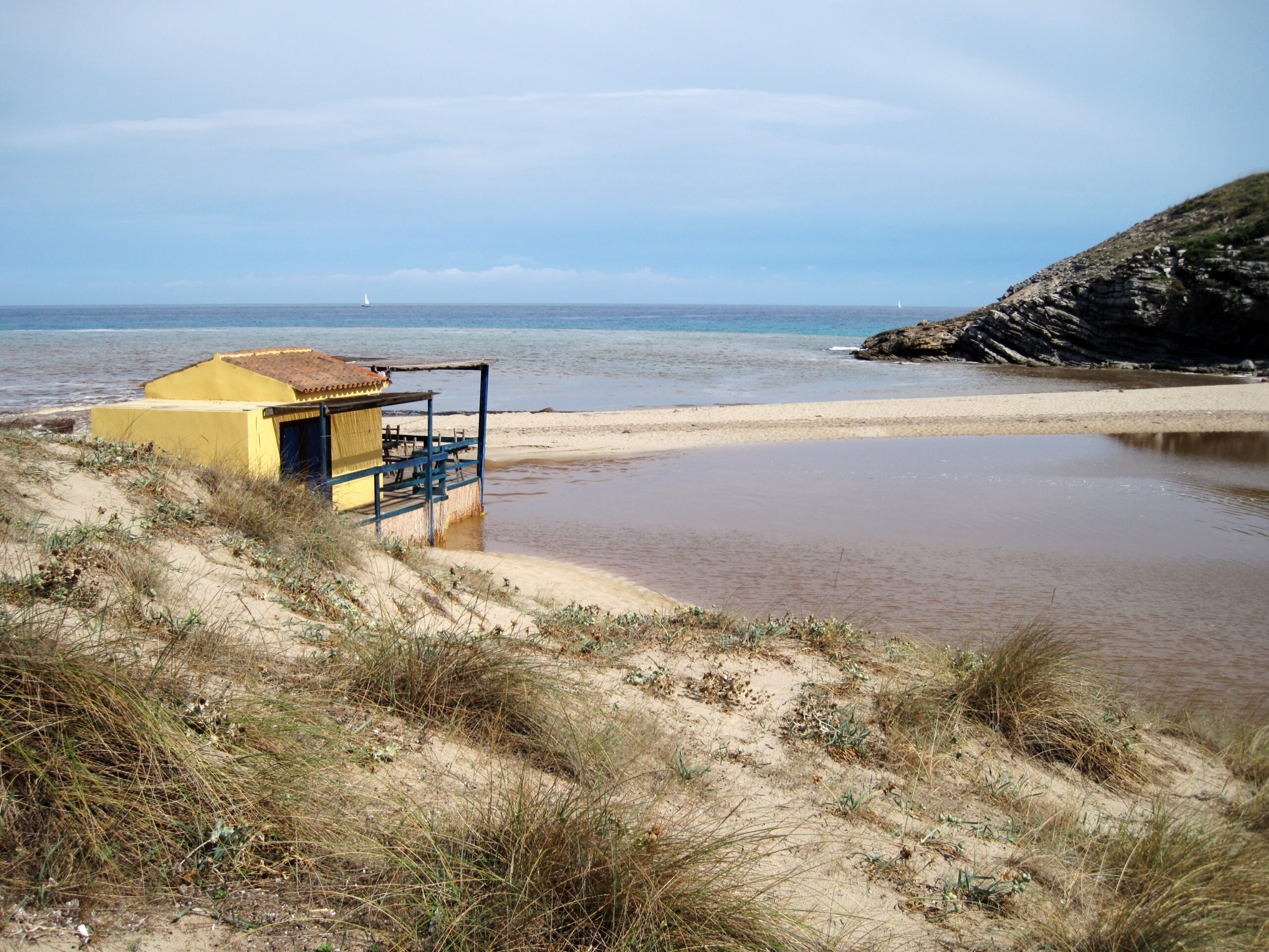 Het gele hutje van de badmeester op de duinrand van Cala Torta, met helmgras op de voorgrond en de brede poel van de torrent naast het strand