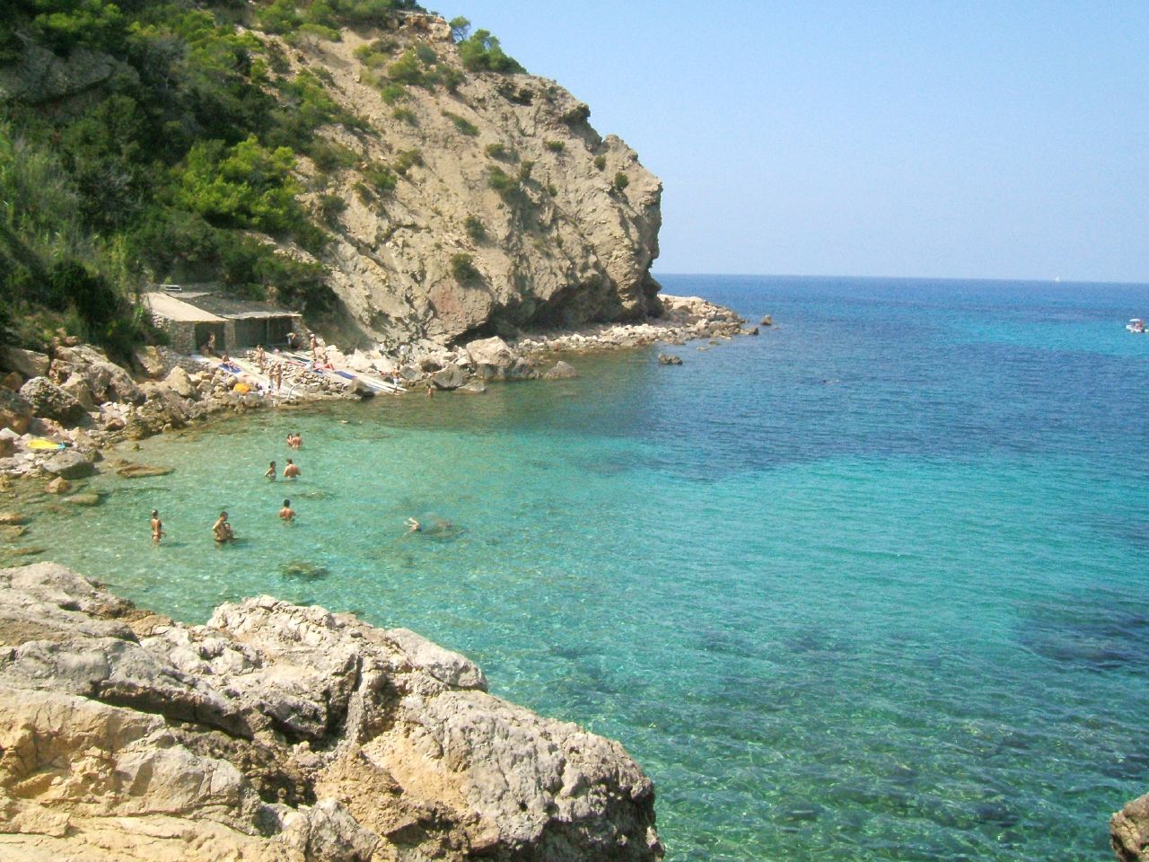 Cala Xarraca van bovenaf gezien, met de steile kalkrots, het lage gebouw van de chiringuito eronder en zwemmers in het ondiepe groenblauwe water