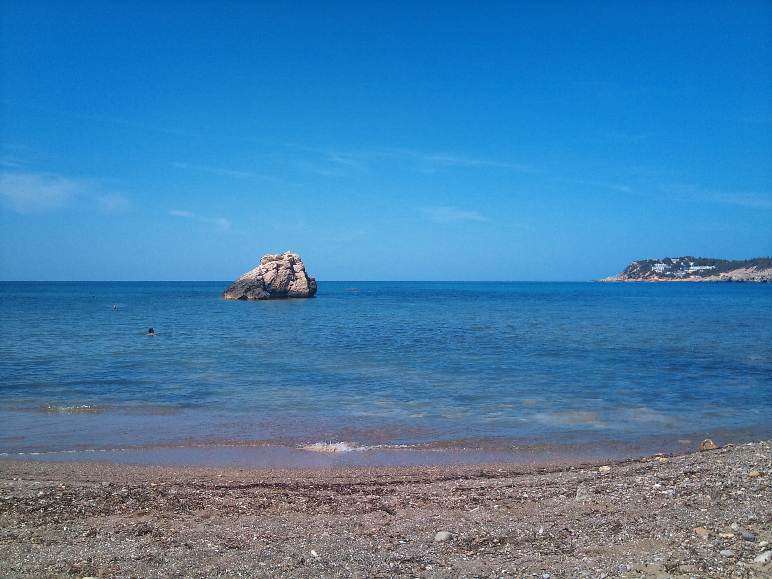 De kiezelstrook van Cala Xarraca bij spiegelglad water, met de losse rotspunt voor de baai en de kaap rechts aan de horizon