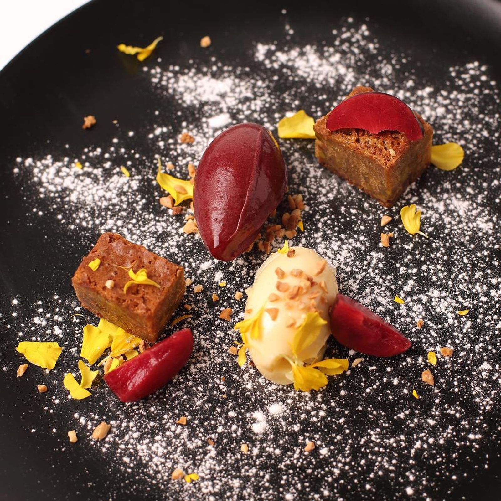 Ca'n Boqueta (Sóller): Bib Gourmand aan de Gran Via 7 Dessert van Ca'n Boqueta met twee sorbetquenelles, blokjes cake en rode saus op een zwart bord met poedersuiker