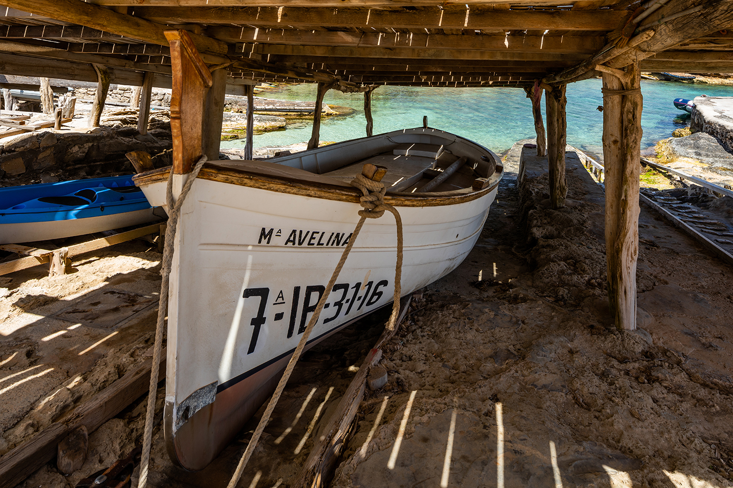 Een llaut op de helling onder een houten escar in het haventje van Es Calo, met het turquoise water erachter