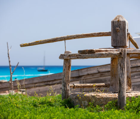 Het oude houten botenhuisgerei bij het haventje van Es Caló de Sant Agustí, met turquoise zee en een zeilboot erachter