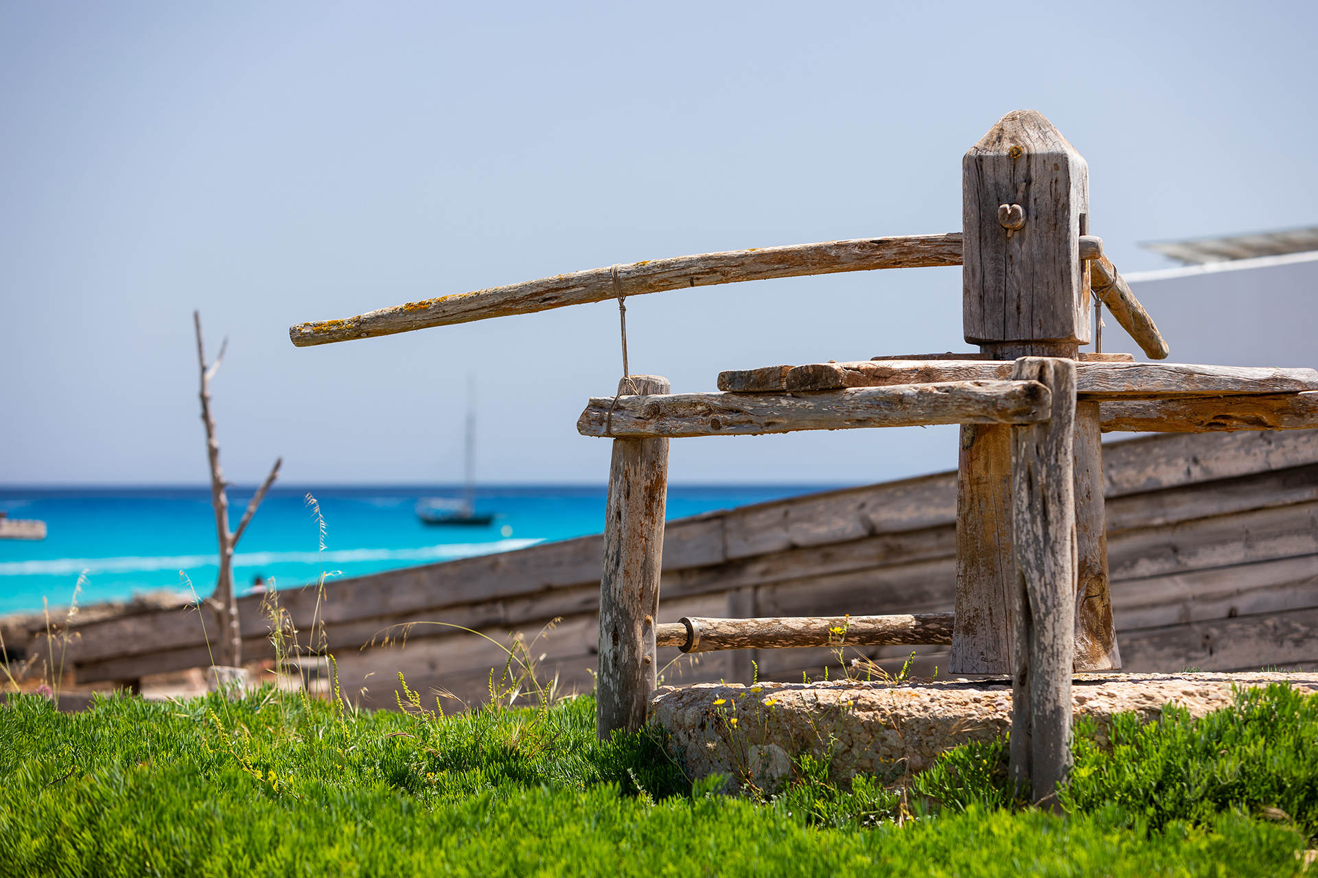 Het oude houten botenhuisgerei bij het haventje van Es Caló de Sant Agustí, met turquoise zee en een zeilboot erachter