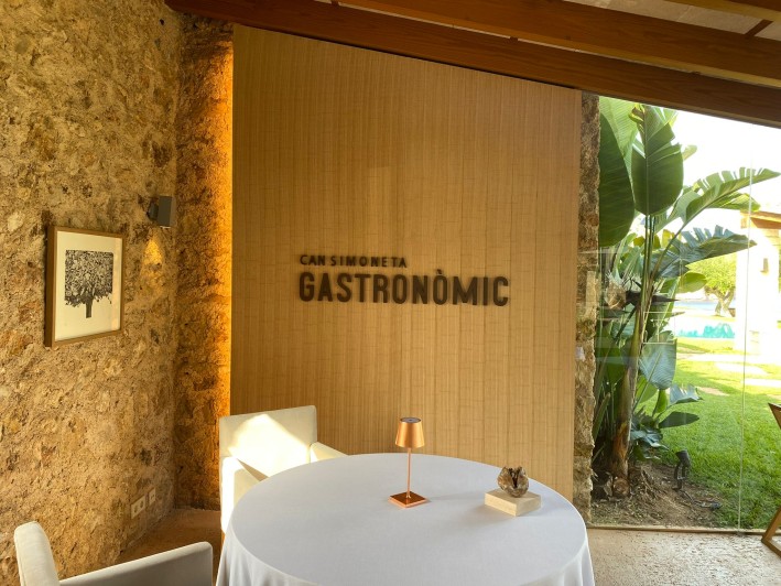 Can Simoneta Gastronòmic (Canyamel): Mexicaans-Mallorcaans menu op de klif 1 De zaal van Can Simoneta Gastronomic met de naam in houten letters op de wand, een ronde gedekte tafel en een natuurstenen muur
