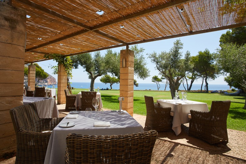Can Simoneta Gastronòmic (Canyamel): Mexicaans-Mallorcaans menu op de klif 8 Het terras van Can Simoneta onder een rieten pergola, met gedekte tafels, olijfbomen en uitzicht over de baai van Canyamel