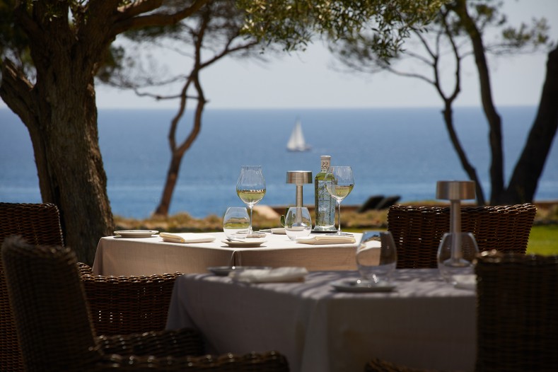 Can Simoneta Gastronòmic (Canyamel): Mexicaans-Mallorcaans menu op de klif 7 Gedekte tafel van Can Simoneta onder de bomen met wijnglazen en uitzicht op de zee met een zeilboot