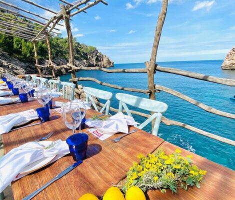 Het terras van Ca's Patró March boven Cala Deià, met een gedekte houten tafel onder een pergola van ruwe boomstammen en het turkooizen water van de baai erachter