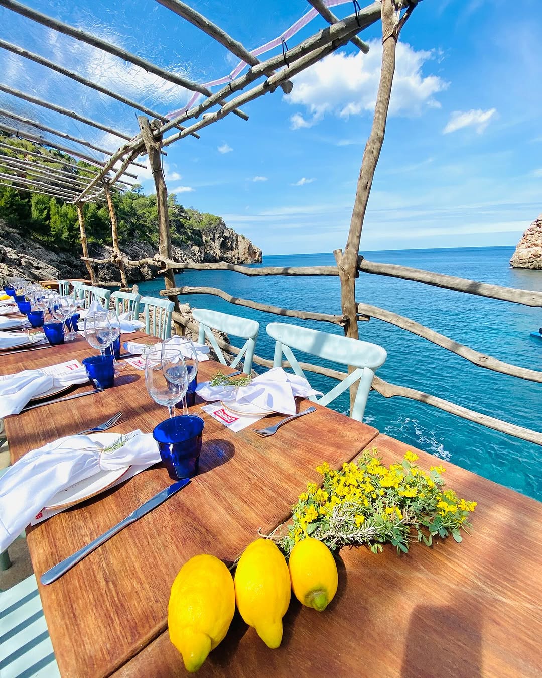 Het terras van Ca's Patró March boven Cala Deià, met een gedekte houten tafel onder een pergola van ruwe boomstammen en het turkooizen water van de baai erachter