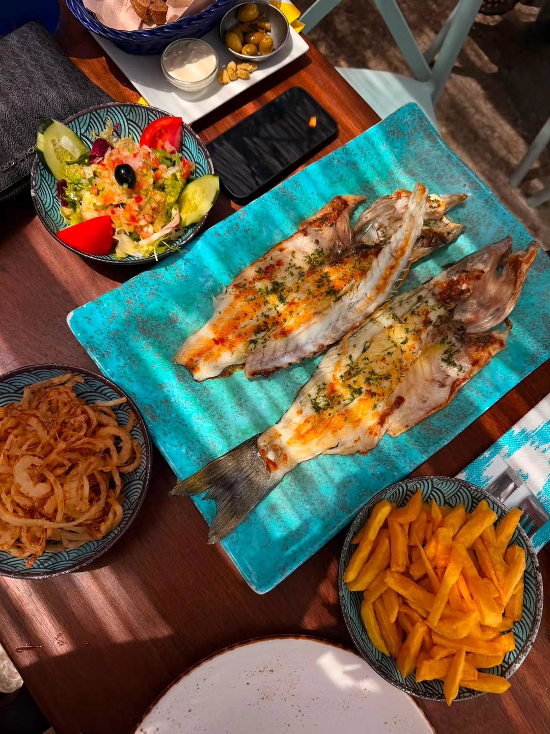 Twee gegrilde vissen op een turkooizen schaal bij Ca's Patró March, met daarnaast schaaltjes friet, gefrituurde ui en een gemengde salade